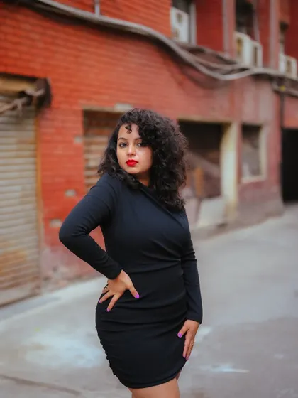 A confident portrait of a model in a black dress against a brick wall. The shallow depth of field and warm tones create a classic urban fashion shot.