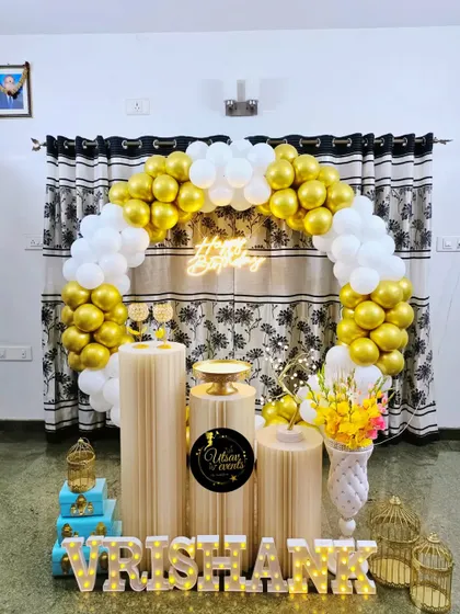 A simple and elegant home birthday decor in white and gold. The balloon arch, neon sign, and plinth arrangement create a clean and classic look.