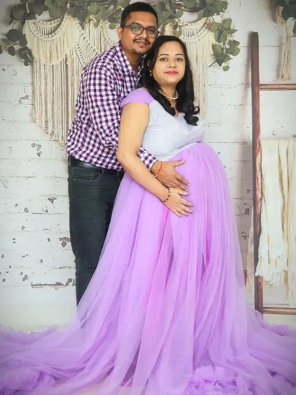 A full-length studio portrait of an expecting couple. This shot showcases the beautiful lilac maternity gown and the supportive connection between the partners.