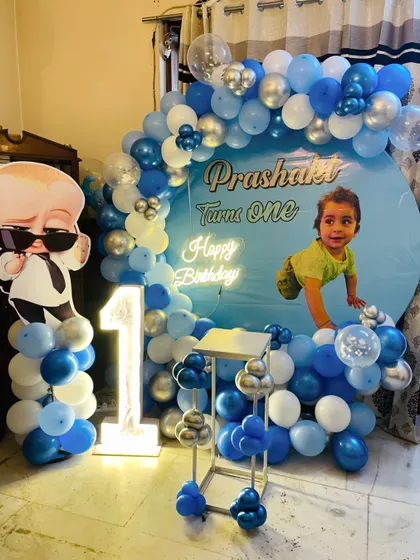 A personalized Boss Baby first birthday. This setup includes a custom backdrop with the child's photo, a blue and white balloon arch, and a light-up number '1'.