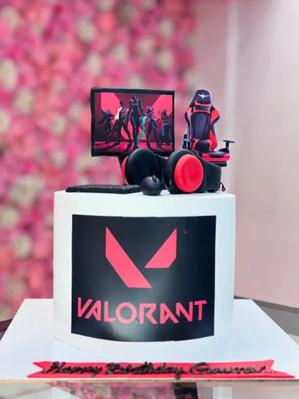 A cake for a Valorant gamer. It features the game's logo and a miniature gaming setup on top, including a monitor, chair, and headset, all made from fondant.