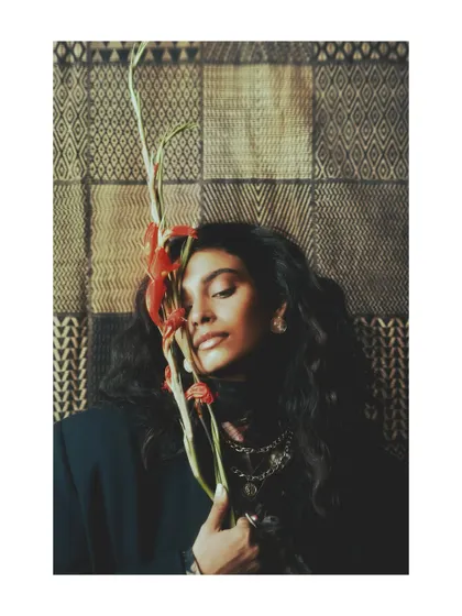 A tender moment from the "Roots" editorial. The subject holds a flower to her face, suggesting a gentle connection to nature and the organic process of growth and self-discovery.