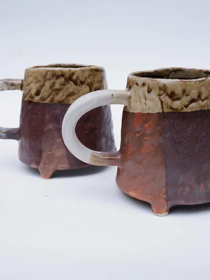 A pair of rustic, three-legged mugs. These are hand-built with a textured, multi-toned glaze and a simple, strong handle. They feel sturdy and comforting to hold.