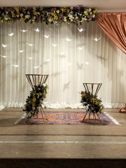 This unique stage design was for a 60th-anniversary celebration. It features a clean white backdrop with hanging bird motifs, asymmetrical floral arrangements, and elegant draping, symbolizing a flight through many years together.