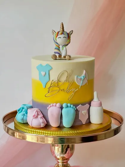A sweet unicorn-themed baby shower cake. The design includes rainbow ombre buttercream and is decorated with baby essentials like booties, bottles, and footprints.
