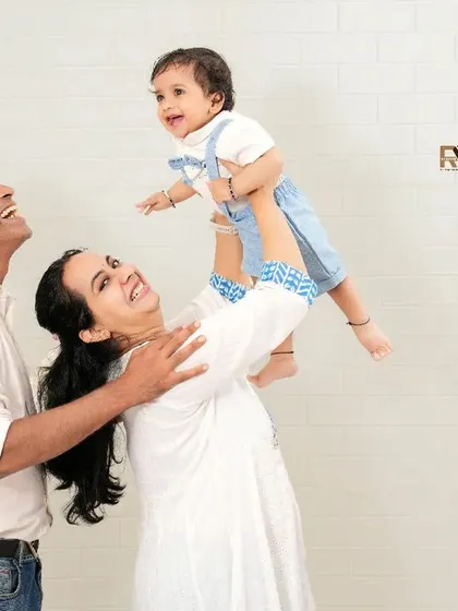 A beautiful family portrait capturing a moment of pure joy. The parents and older sister look lovingly at the baby being lifted in the air, creating a dynamic and happy image.