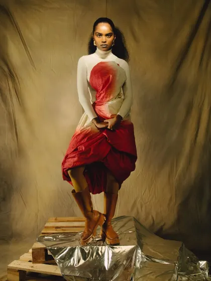 A full-body studio portrait featuring a unique, hand-painted dress. The model's strong pose on a silver-draped platform showcases the garment as a piece of wearable art.
