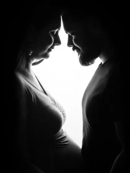A dramatic silhouette of an expecting couple. The high-contrast black and white lighting creates an intimate and artistic portrait focused on their connection.