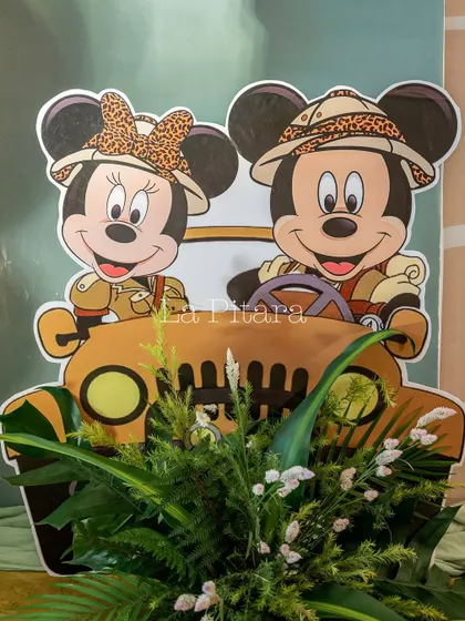A cute cutout of Mickey and Minnie in a safari jeep, used as a decorative element within a floral arrangement for a jungle-themed party.