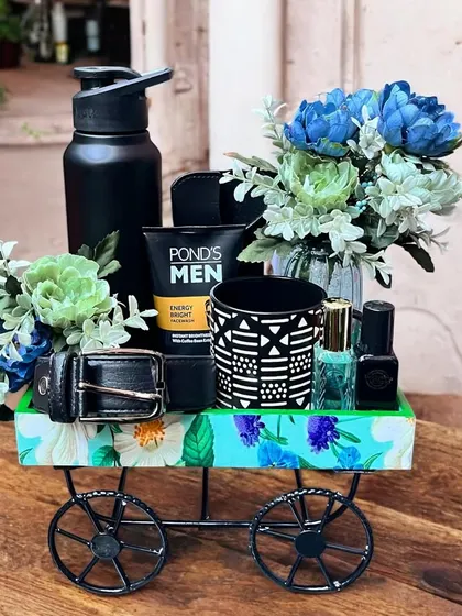 This creative men's gift hamper is assembled on a decorative cart. It's filled with grooming essentials, a water bottle, a belt, and a small floral arrangement for a stylish presentation.