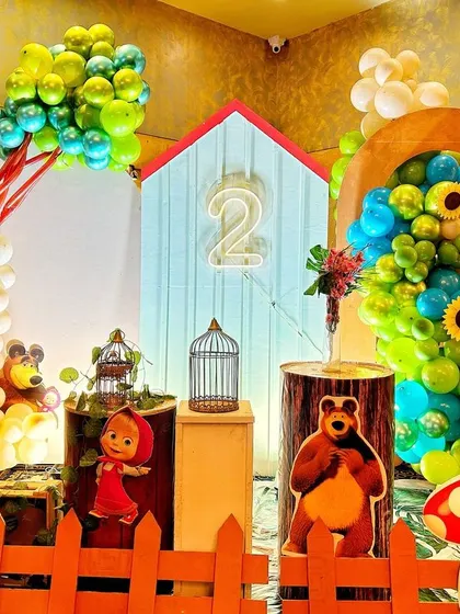 A delightful Masha and the Bear themed birthday setup. The decor features a house-like backdrop, character cutouts, toadstool props, and balloon arrangements that look like trees.