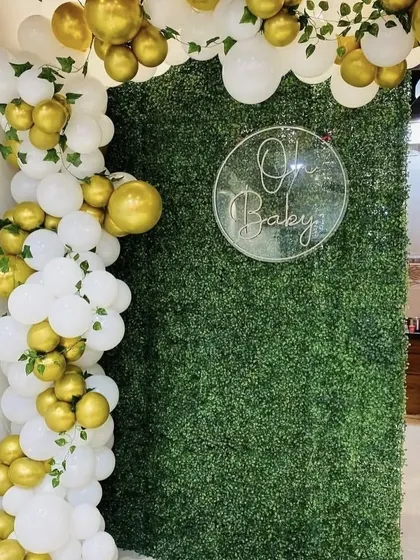 An elegant baby shower backdrop using a lush green foliage wall. The white and gold balloon garland with ivy accents creates a fresh and sophisticated look.