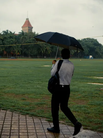 A man in formal wear walks along a paved path in a park, holding an umbrella. It's a slice of the daily commute for many Mumbaikars during the rainy season.