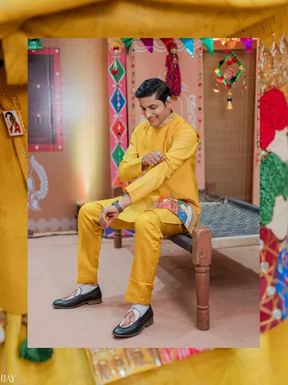 The groom adjusts his sleeve, a candid moment captured against the colorful, rustic Haldi backdrop.