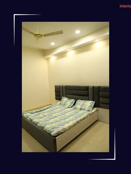 A modern bedroom with a custom grey upholstered headboard. The false ceiling incorporates cove lighting, which provides a soft, indirect glow.