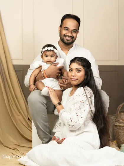 A classic family portrait in all-white attire. The baby sits on her father's lap while her mother sits gracefully on the floor, creating a balanced and serene image.