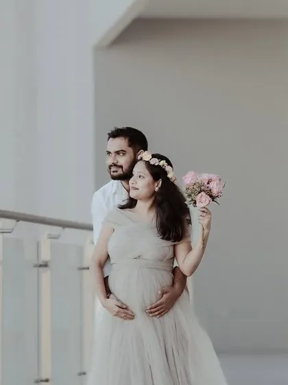 A tender moment between an expecting couple on a balcony. The partner holds his wife from behind as she holds a bouquet, both looking towards the future.