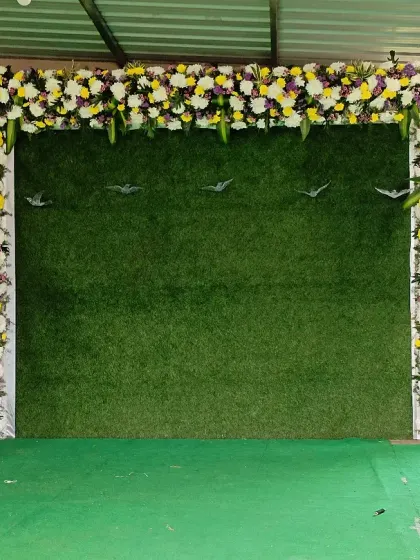 This simple yet stylish backdrop is perfect for photos at any event. It features an artificial grass wall framed by fresh flowers, flanked by two ornate golden vases with matching floral arrangements.