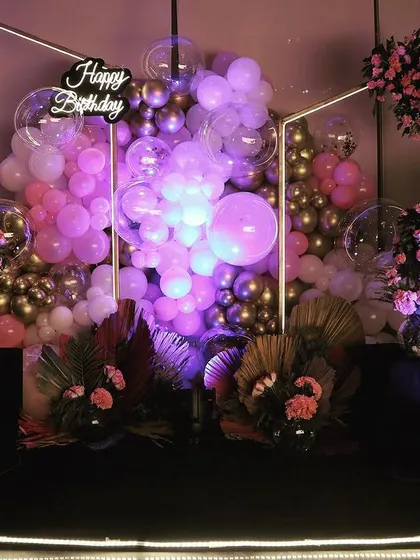 A birthday party photo backdrop featuring a geometric frame, a cascade of pink and purple balloons, and dramatic uplighting for a fun and glamorous feel.