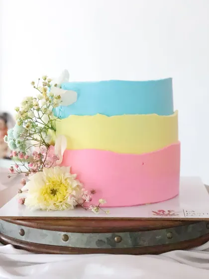 A colorful pastel cake with layers of pink, yellow, and blue. The raw edge and fresh flower accents give it a modern and cheerful look.