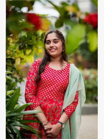 A smiling portrait of a young woman in a vibrant red dress, captured with natural light in a garden setting.
