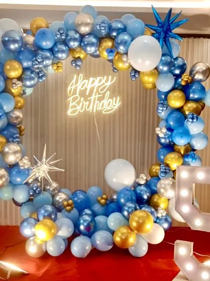 A 5th birthday setup with a circular balloon frame in shades of blue, silver, and gold. The decor includes a "Happy Birthday" neon sign and a marquee number '5'.