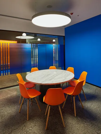 A round meeting table with vibrant orange chairs set against a deep blue wall and a fluted glass partition. The bold color contrast makes this a visually dynamic space.