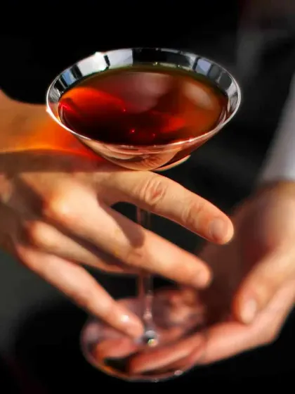 A bartender holding a classic coupe glass with a dark, spirit-forward cocktail. This showcases our expertise in crafting timeless drinks like the Manhattan or Negroni.