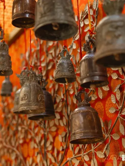 Antique temple bells hang against a vibrant orange backdrop, echoing the sounds and sights of a sacred sanctum. These details are essential to creating the immersive 'Divya Stambh' experience.