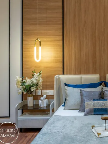 This bedroom corner showcases a beautiful blend of textures. A wood-paneled accent wall is paired with a cream-colored headboard and pillows in shades of blue. The modern pendant light and white nightstand with floral decor complete this stylish and comfortable setup.