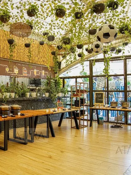 An indoor venue transformed for a soccer-themed party. The space features long wooden tables, a ceiling draped with greenery and soccer balls, and a buffet station, ready for a team celebration.