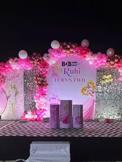 A glamorous Barbie-themed stage for a second birthday. The outdoor setup includes a large stage, multiple backdrops with Barbie illustrations, a shimmer panel, and a pink balloon arch.