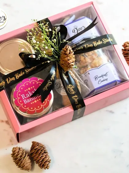 A pretty pink gift box curated for Rakhi. This hamper includes Breakfast Cookies and other treats, decorated with a black ribbon and natural pinecones for a rustic touch.