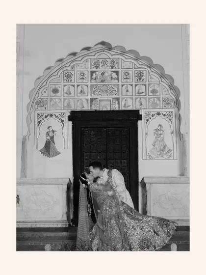 A romantic black-and-white photo of the groom kissing the bride's forehead in front of an ornate, traditional doorway.