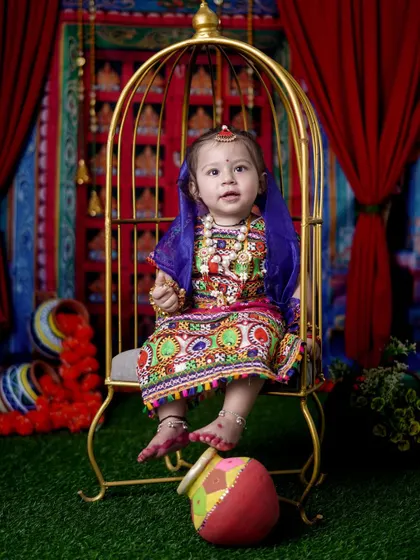 Here is a little Radha in a stunning, colorful lehenga, seated in a golden birdcage prop. The vibrant backdrop and traditional matki complete the look for this festive portrait.