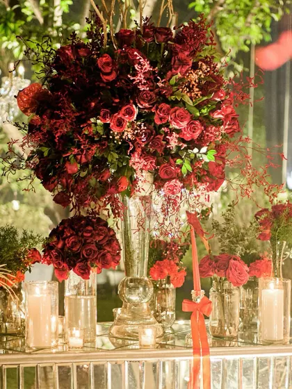 A close-up of a lush centerpiece, showing the mix of deep red roses and other textured florals. The arrangement is reflected in the mirrored table, doubling its impact.