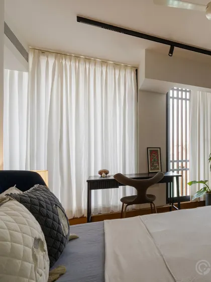 This bedroom corner is designed as a functional workspace, with a simple desk and chair positioned to receive ample natural light. Sheer white curtains diffuse the light, creating a bright yet private area.