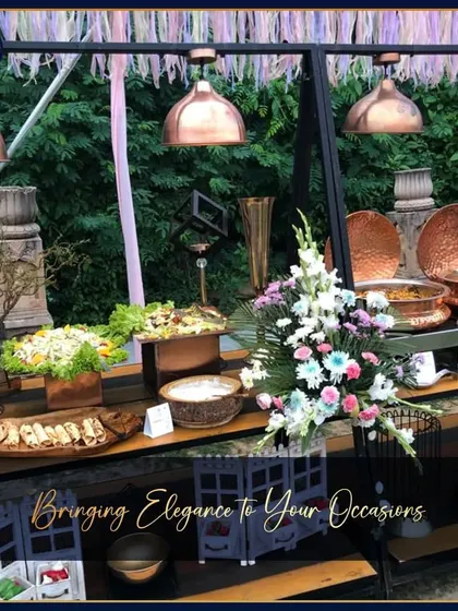 An outdoor buffet station with a rustic-chic aesthetic. The setup features copper lamps, wooden platters, and abundant floral arrangements, creating a warm and inviting atmosphere.