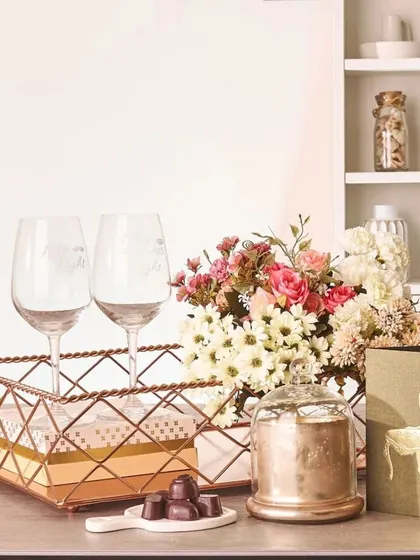 A romantic and elegant gift set, this arrangement features two wine glasses in a rose gold wire basket, a bouquet of fresh flowers, premium chocolates, and a decorative candle.