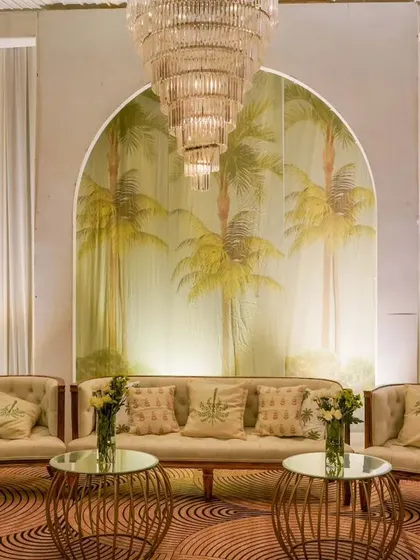 A chic lounge area designed for a reception, featuring a tropical-themed mural as a backdrop. The neutral-toned furniture and gold accents create a relaxed yet sophisticated space for guests to network and socialize.