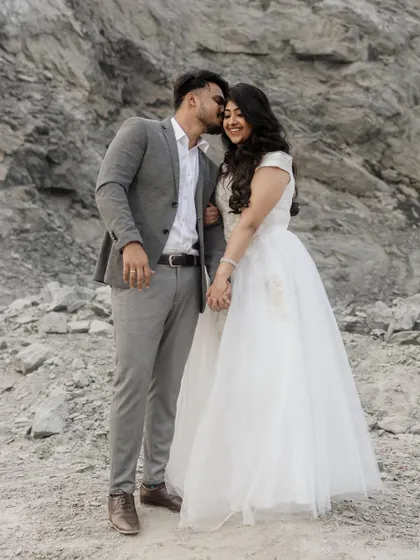 A sweet and affectionate moment against a rugged, natural backdrop. He kisses her on the cheek as she smiles, holding his hand. This full-length shot captures their joy and the beautiful contrast of her white dress against the rocks.