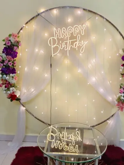 A simple and elegant birthday ring with fairy lights, white drapes, and artificial flower accents. The neon sign is reflected on the glass table below.