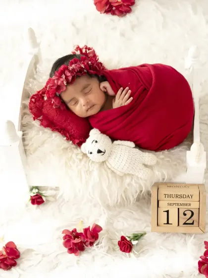 A beautiful newborn girl in a red wrap and floral headband, sleeping on a white bed with a calendar block marking the date.