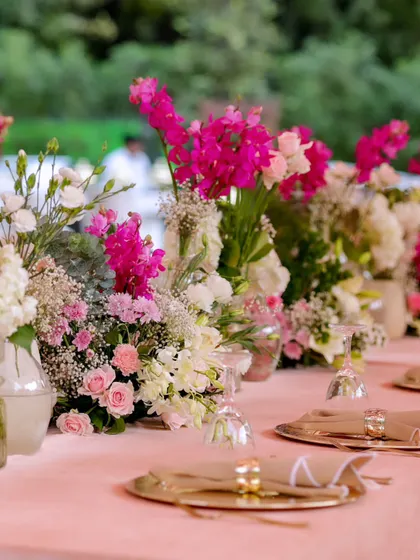 A beautifully set dining table with a lush runner of pink and white flowers, complemented by gold charger plates and elegant glassware for a refined dining experience.