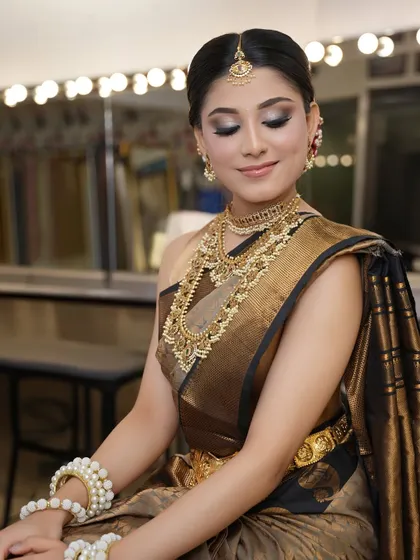 The bride seated, showcasing her complete look. The traditional hairstyle and makeup are designed to complement the classic silk saree and gold jewelry perfectly.