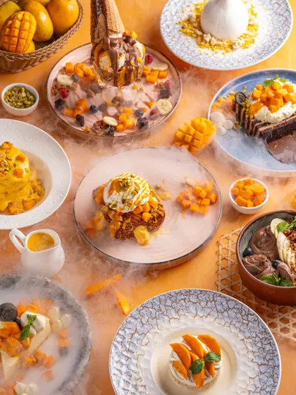 A stunning top-down view of the mango dessert festival at Bastian. The image is a feast for the eyes, showcasing a dozen different creations with dramatic dry ice smoke.