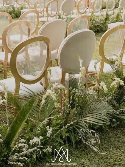 Elegant transparent chairs with gold frames line the aisle, which is flanked by lush, low-profile greenery arrangements, leading the eye towards the sea.
