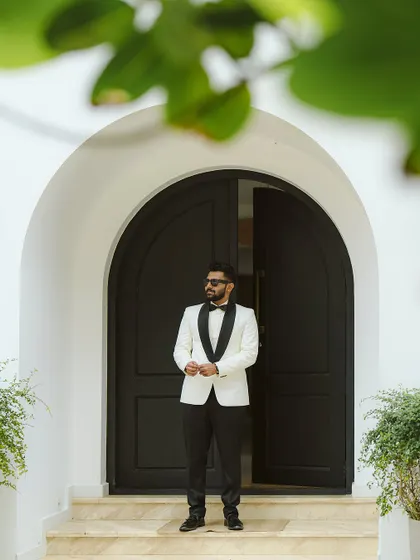 A groom in a stylish white tuxedo jacket stands in a grand arched doorway. This portrait exudes modern sophistication and confidence.