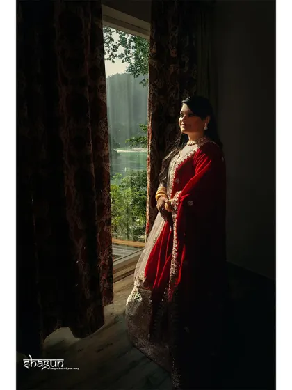 In the spotlight of love. The bride stands by a window, the natural light creating a beautiful silhouette and highlighting her red and white lehenga.