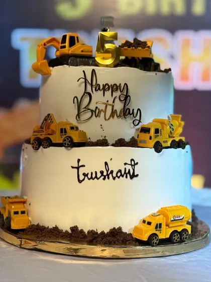 A full view of the two-tier construction cake, showing all the different toy trucks and the personalized name 'Trushant'.